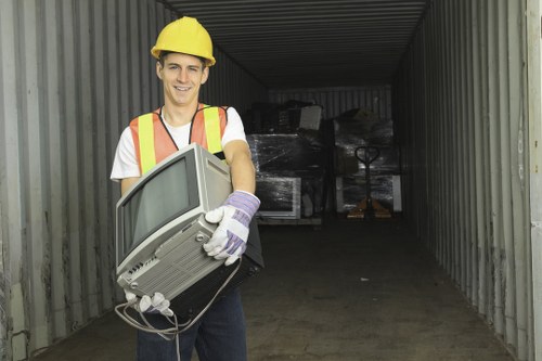 A young man dressed in a yellow safety helmet, high-visibility vest, and work gloves is standing inside a large shipping container with metal walls and ceiling. He is smiling and holding a small, older model CRT television in his hands, with the screen facing outward. Behind him, the container's interior appears dark and contains a mixture of bulky rubbish, including black plastic-wrapped items, which are stacked irregularly towards the rear. The container is situated outdoors, possibly on a driveway or service yard, and the surrounding environment is not visible in the image. The scene reflects a rubbish removal service context, with the young worker preparing to transport or clear unwanted items, aligning with house clearance activities typical in Radlett and nearby areas.