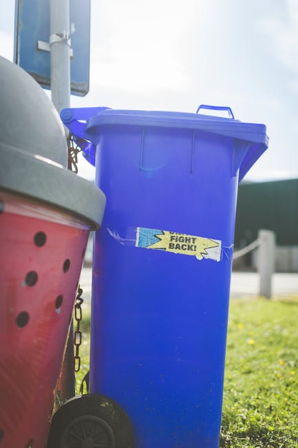 A black, classic-style street rubbish bin with a rounded, domed lid and vertical ribbed design sits on a cobblestone pavement in an urban environment. The top of the bin is cluttered with various disc
