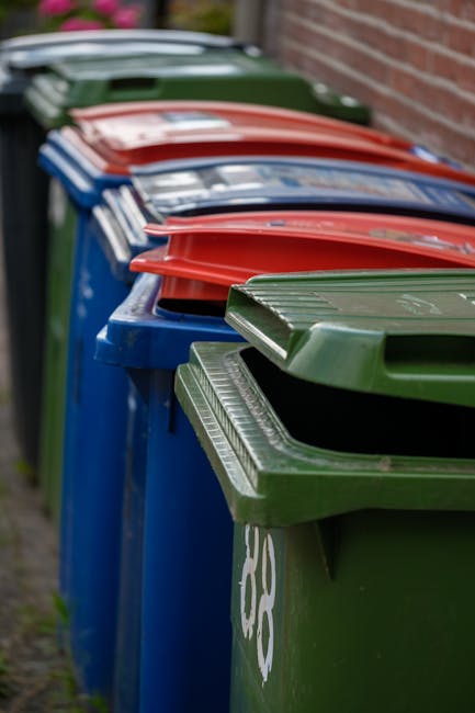 A row of large wheelie bins positioned along a pavement against a brick wall, used for waste collection. The bins are made of plastic, with textured lids featuring ridges for stacking, and are painted